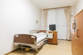 介護施設の個室。電動ベッド、テレビ、木製家具、明るい窓が配置されている。
